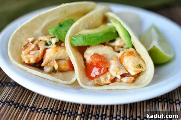 Seared Cilantro Lime Fish Tacos 2 Two fish tacos in corn tortillas with avocado slices and diced tomatoes on a white plate, ready to be enjoyed.