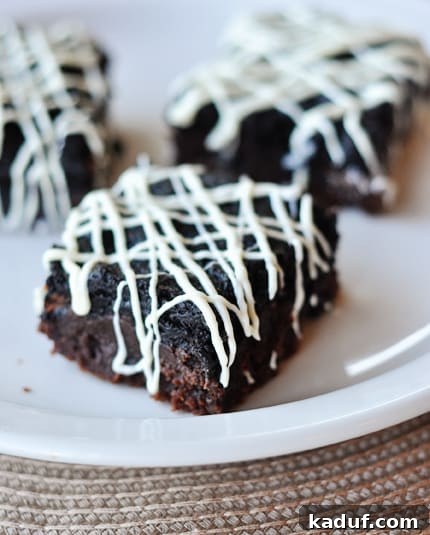 Oreo Truffle Fudge Brownies 3 Three decadent Oreo truffle brownies, each adorned with a white chocolate drizzle, arranged artfully on a white plate, showcasing their rich layers.