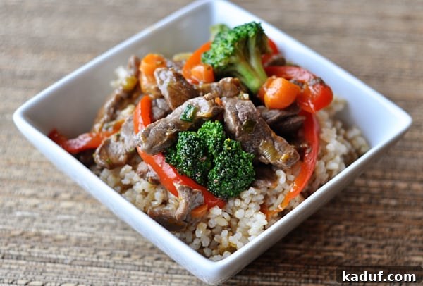 Zesty Orange Beef & Vegetable Stir-Fry 3 Close-up of a square white bowl showcasing cooked rice adorned with vibrant broccoli, red peppers, and savory beef strips.