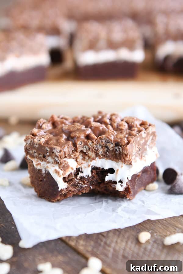 Ultimate Crunch Brownies 2 Killer crunch brownie on white paper with a bite taken out, showcasing the distinct layers of fudgy brownie, marshmallow, and crispy peanut butter chocolate topping.