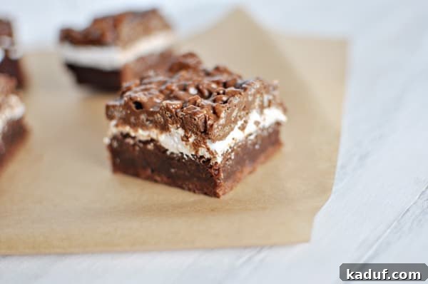 Ultimate Crunch Brownies 12 Close-up of a brownie with a distinct brownie bottom, a gooey marshmallow middle, and a crunchy chocolate peanut butter topping, cut into a small square on white parchment paper.