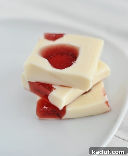 Stained Glass Heart Treats 3 Three layered squares of white Jello with vibrant red Jello hearts inside, artfully stacked on a white plate, ready to be enjoyed.