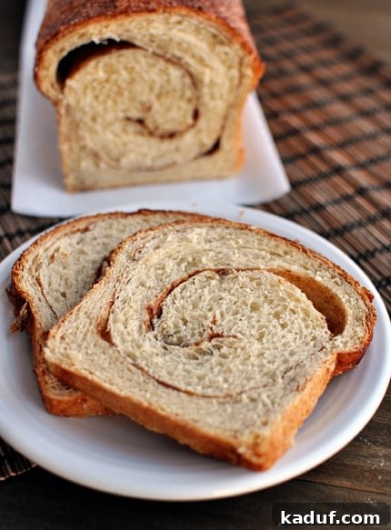 Ultimate Cinnamon Swirl Loaf 3 Two golden-brown slices of cinnamon swirl bread on a white plate, with the rest of the beautifully swirled loaf blurred in the background, inviting a delicious breakfast.