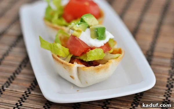 Tiny Taco Treats 4 Two vibrant mini taco cups, generously topped with fresh lettuce, diced tomatoes, a dollop of sour cream, and creamy avocado, arranged on a pristine white platter.