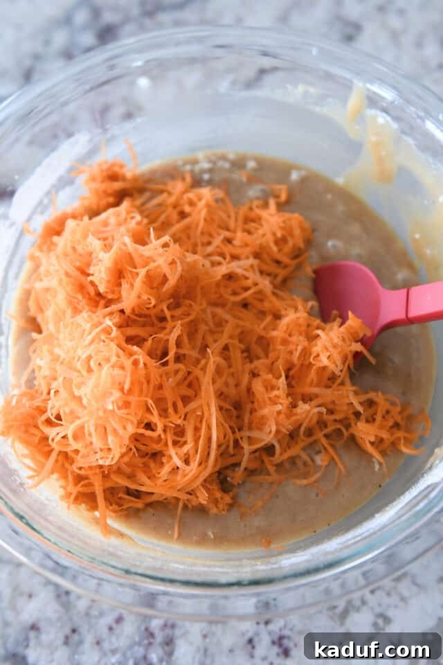 Carrot Cake Cheesecake Fusion 5 Finely shredded carrots, vibrant orange, mixed into the golden cake batter in a clear glass bowl, ready for baking.