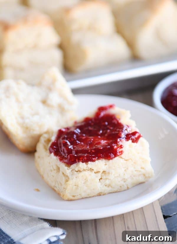 Perfectly Flaky Buttermilk Biscuits 4 A flaky buttermilk biscuit cut in half on a white plate, served with a dollop of jam and a pat of butter.