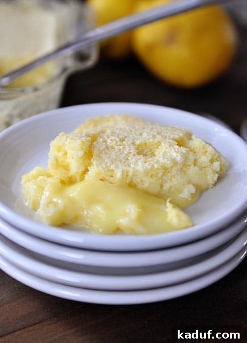 Zesty Lemon Pudding Cake 2 A slice of vibrant Lemon Pudding Cake on a white plate, showcasing its distinct two layers: a golden, airy cake on top and a creamy, yellow pudding layer underneath. The cake is garnished with a sprig of mint and a dusting of powdered sugar, with a stack of clean white plates in the background.