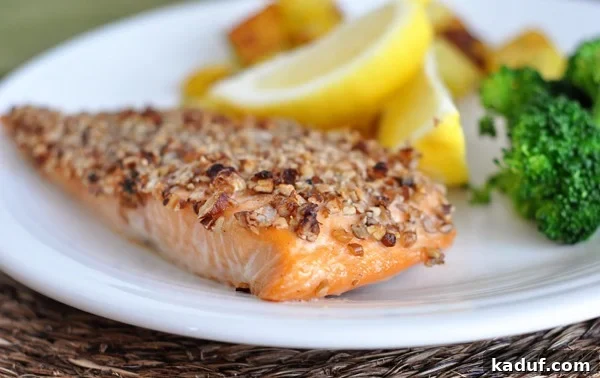Maple Pecan Glazed Salmon 2 Pecan-crusted salmon fillet on a white plate, garnished with fresh lemon slices and a side of steamed broccoli. The salmon is golden-brown with a visible pecan crust.