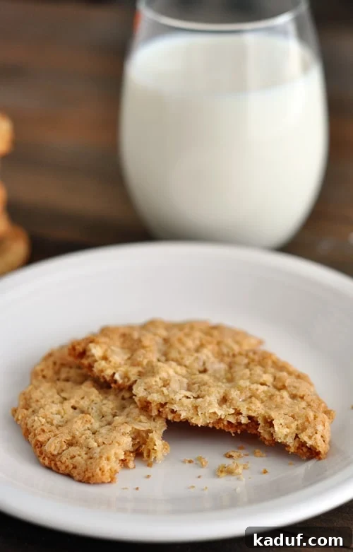 Chewy Oatmeal Lace Cookies 8 A thin, baked oatmeal cookie split in half on a white plate, with a glass of milk in the background.