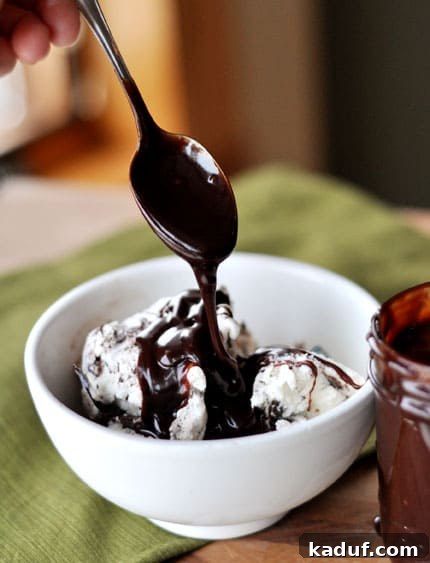 Velvety Warm Chocolate Topping 2 A white bowl of ice cream with a spoon elegantly drizzling rich, homemade hot fudge sauce on top, highlighting its luscious texture.