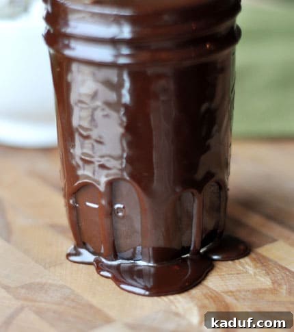 Velvety Warm Chocolate Topping 3 A charming glass jar filled to the brim with glossy hot fudge sauce, with some deliciously spilling over the sides, ready to be enjoyed.