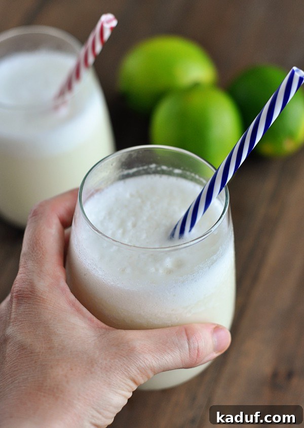 Velvet Brazilian Lemonade 2 A tall glass of frothy, creamy Brazilian Lemonade (limeade) with a blue and white striped straw, garnished with fresh lime slices in the background, set against a bright, inviting backdrop.