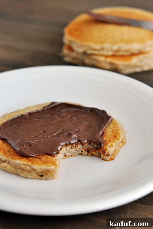 Easy Overnight Cinnamon Oat Pancakes 2 A pancake topped with Nutella, with a bite taken out, on a white plate.