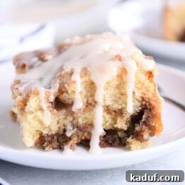 Square of cinnamon roll cake on white plate with glaze on top and sides.