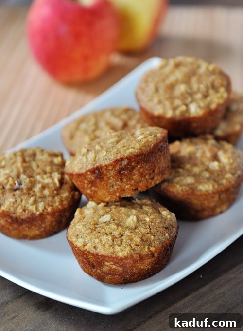 Wholesome Apple Oat Muffins 7 A white platter generously piled with freshly baked applesauce oat muffins, their tops golden and inviting. Whole apples and cinnamon sticks blur softly in the background, hinting at the key flavors.