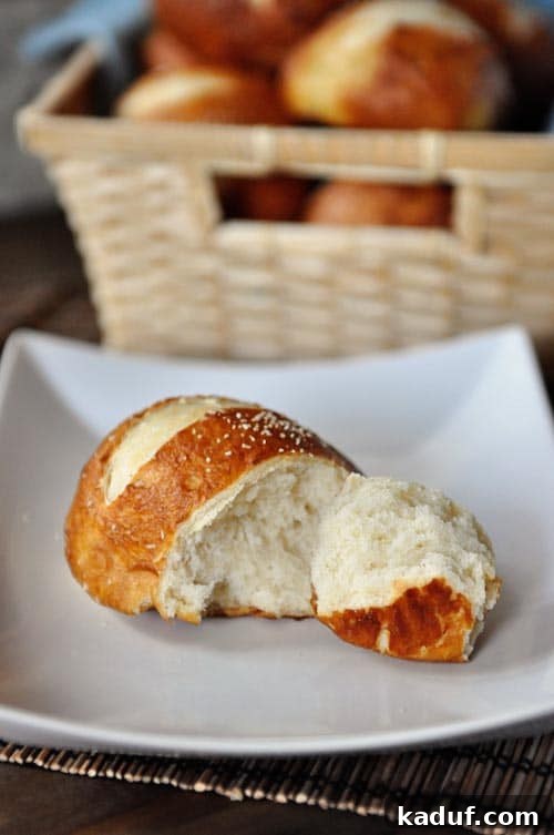 Chewy Pretzel Buns 4 A golden brown pretzel roll with a chunk split off on a white plate, highlighting its chewy interior.