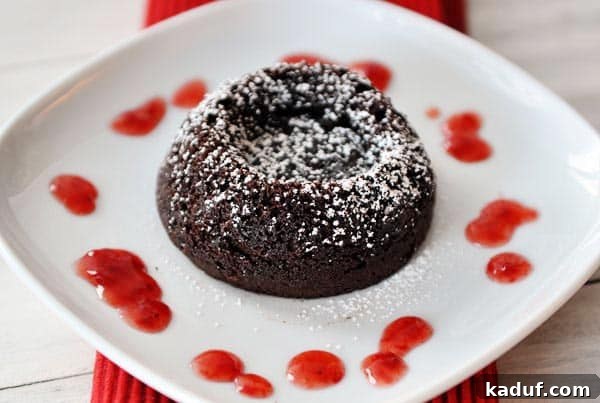 Guilt-Free Molten Chocolate Fudge Cakes 2 A perfectly baked chocolate molten cake, dusted with powdered sugar and drizzled with vibrant raspberry sauce, sits on a pristine white plate, promising a decadent experience.