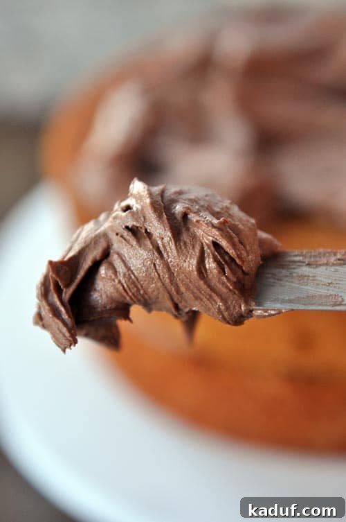 Decadent Chocolate Cloud Frosting 3 A metal spatula with creamy chocolate frosting, with a blurred background of a cake being frosted, highlighting the smooth texture.