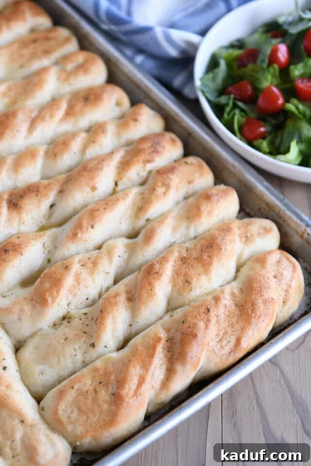 Heavenly 60-Minute Homemade Breadsticks 7 A vibrant green salad sits in the background, out of focus, while a pan of freshly baked, golden breadsticks takes center stage, ready to be enjoyed.