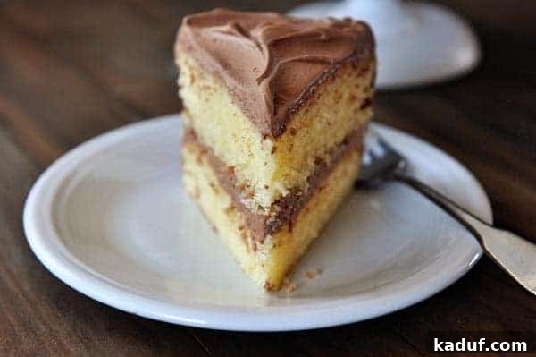 The Gold Standard Homemade Yellow Cake 12 A perfect slice of tender yellow cake with rich chocolate frosting, gracefully placed on a white plate with a fork poised beside it, inviting a delicious bite.