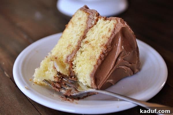 The Gold Standard Homemade Yellow Cake 4 A fork taking a bite out of a delightful piece of fluffy yellow cake topped with rich chocolate frosting on a pristine white plate.