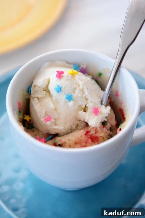 Vegan Vanilla Funfetti Mug Cake 2 A white mug with a cooked funfetti mug cake, topped with a scoop of vanilla ice cream, sprinkles, and a spoon, ready to be enjoyed.