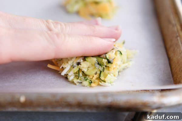 Cheesy Zucchini Gems 6 A hand patting down a cheesy uncooked zucchini bite on a cookie sheet.
