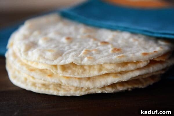 Step-by-Step Guide to Homemade White and Whole Wheat Tortillas 2 A stack of golden brown homemade cooked tortillas rests under a soft blue napkin, hinting at their warmth and fresh texture.