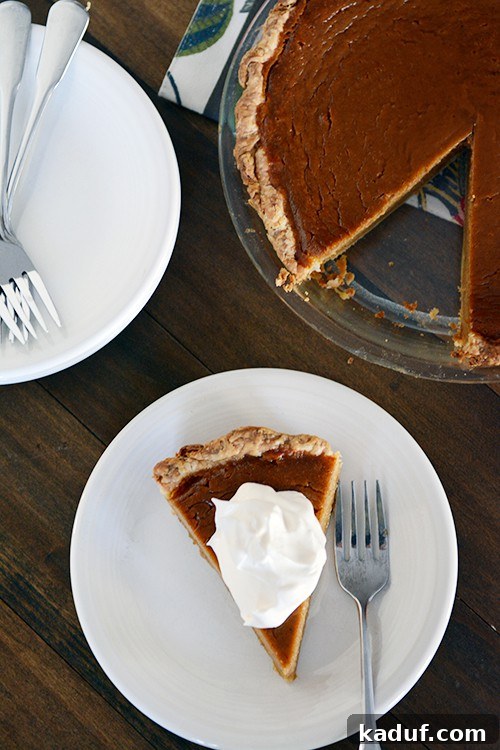 Simply Perfect Pumpkin Pie 2 Top view of a beautifully baked classic pumpkin pie with a slice missing, featuring a dollop of whipped cream on the removed slice on a white plate.
