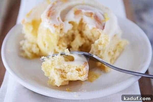 Gooey Lemon Rolls with Zesty Cream Cheese Glaze 3 Frosted sticky bun on white plate with fork taking a bite.