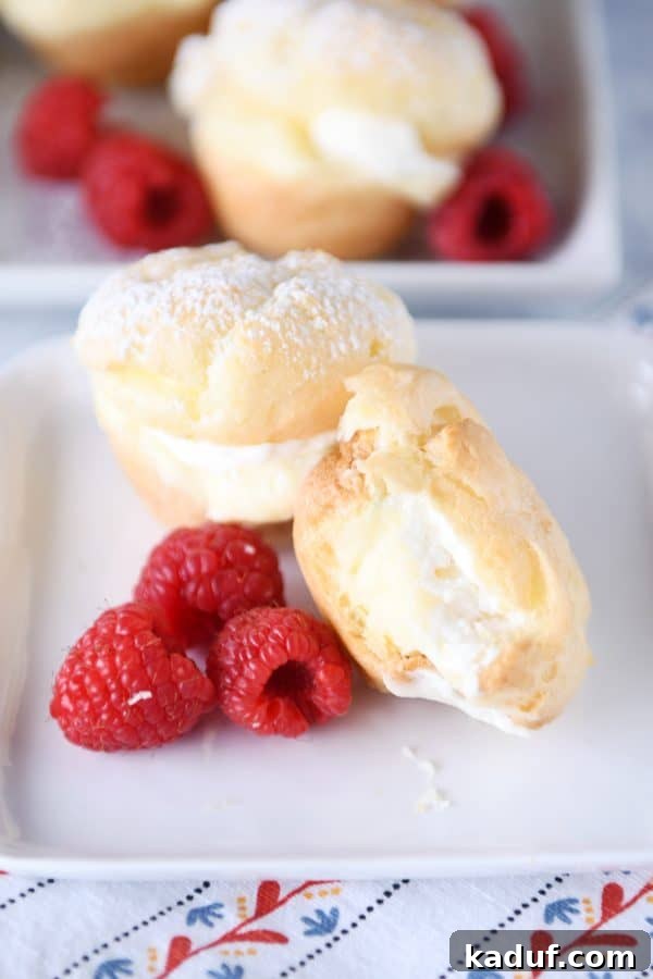 Effortless Cream Puff Perfection 6 A close-up of a mini cream puff on a white plate with raspberries, showing a bite taken out, revealing the creamy filling inside.