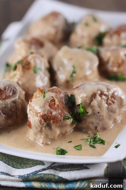 Creamy Skillet Swedish Meatballs 12 A beautifully presented white platter showcases a generous serving of golden brown, perfectly cooked Swedish meatballs, lavishly coated in a gleaming, rich brown gravy.