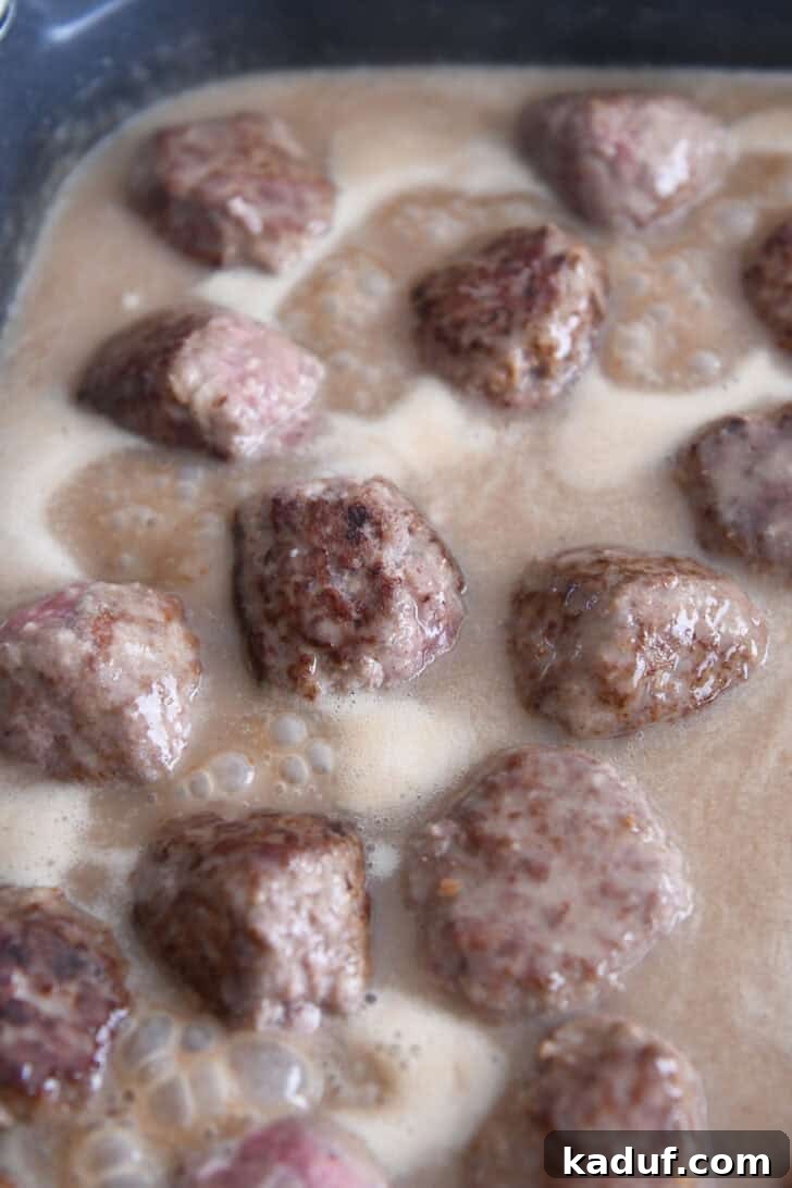 Creamy Skillet Swedish Meatballs 7 Golden-brown Swedish meatballs gently simmering in a luxurious, thick brown gravy sauce within an electric skillet, absorbing all the rich flavors.