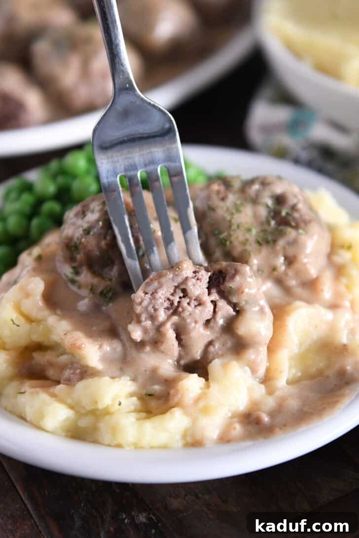 Creamy Skillet Swedish Meatballs 10 A fork gently pierces a tender half meatball, nestled atop a mound of creamy mashed potatoes and generously coated in a luscious brown gravy, showcasing its delicious texture.