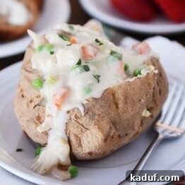 A baked potato with creamy chicken pot pie filling and a side of green peas, garnished with fresh herbs.