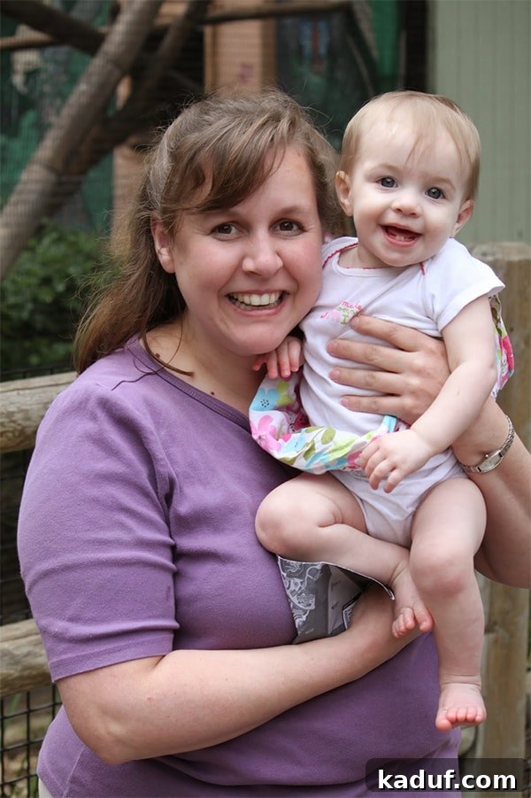 A woman with dark hair, wearing a casual top, holding a baby girl with a pink bow in her hair. They are both smiling.