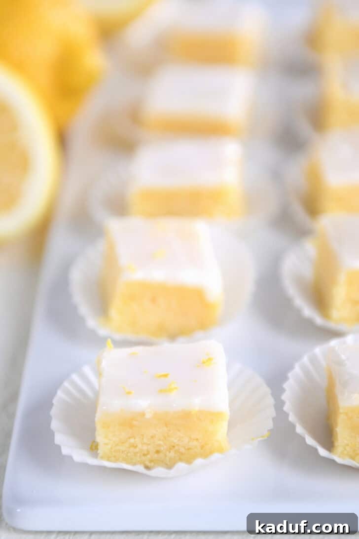 Mini Lemon Brownie Delights 2 A line of lemon brownie squares topped with lemon zest and a white glaze in white muffin cups on a white serving platter, showcasing their delightful appearance and bright, zesty appeal.