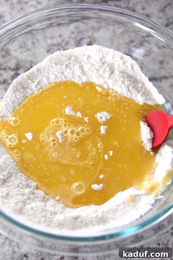Mini Lemon Brownie Delights 3 Top down view of a glass bowl with flour, a lemon, and an egg mixture in the middle of the flour, with a red spatula on the side, demonstrating the initial mixing steps for the lemon brownie batter.