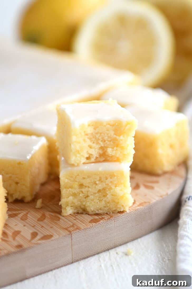 Mini Lemon Brownie Delights 8 Two small lemon brownie squares with a white glaze topping stacked on top of each other on a wooden cutting board. More lemon brownie squares are in the background, creating an inviting rustic scene.