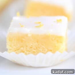 A lemon brownie square topped with a white glaze and lemon zest sitting in a white muffin liner, ready to be served, capturing its delightful presentation.