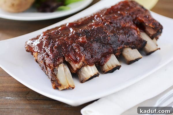 Root Beer Glazed Slow Cooker Baby Back Ribs 5 A freshly broiled rack of slow cooker baby back ribs, glistening with a caramelized root beer BBQ sauce, presented on a pristine white platter.