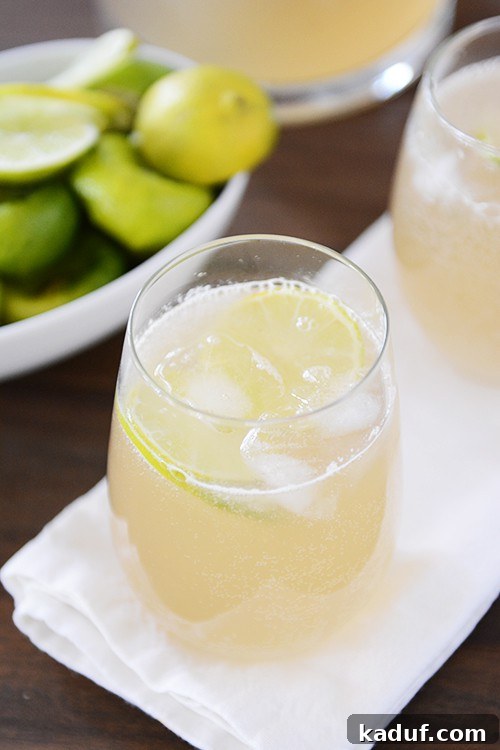 Ginger Mint Lime Fizz 2 Two tall glasses of iced ginger mint limeade, garnished with fresh lime slices and mint sprigs, set against a bright, airy background with a bowl of whole and sliced limes.