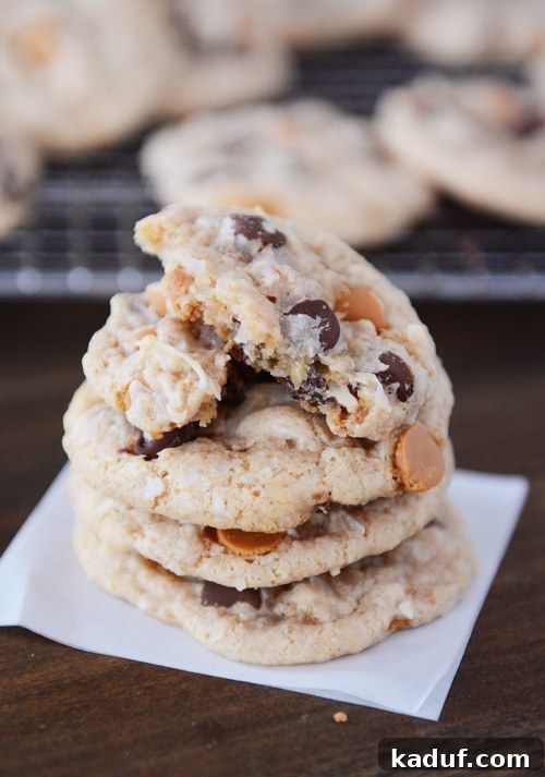 Soft Chewy Seven Layer Cookies Individual Perfection 2 A stack of golden brown 7-layer cookies, featuring melted chocolate and butterscotch chips, with the top cookie expertly broken in half to reveal its soft, chewy interior.