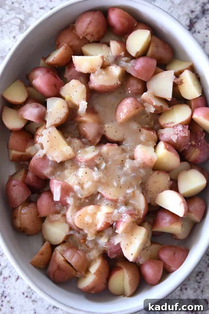 Rustic German Potato Salad 7 Top down view of a white oval dish with large potato chunks and an onion sauce spread over the top, showing the initial mixing of dressing with potatoes.