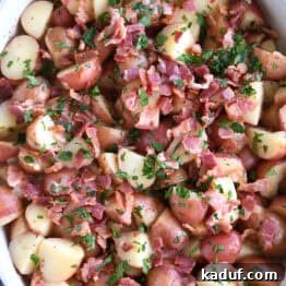 Rustic German Potato Salad 10 Top down view of a white oval dish with potato chunks, crispy bacon pieces, and fresh chopped parsley. A delicious serving of warm German Potato Salad.