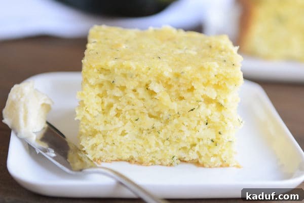 Golden Zucchini Cornbread 4 A thick piece of fluffy zucchini cornbread on a white plate with a knife and butter on the side, ready to be enjoyed.
