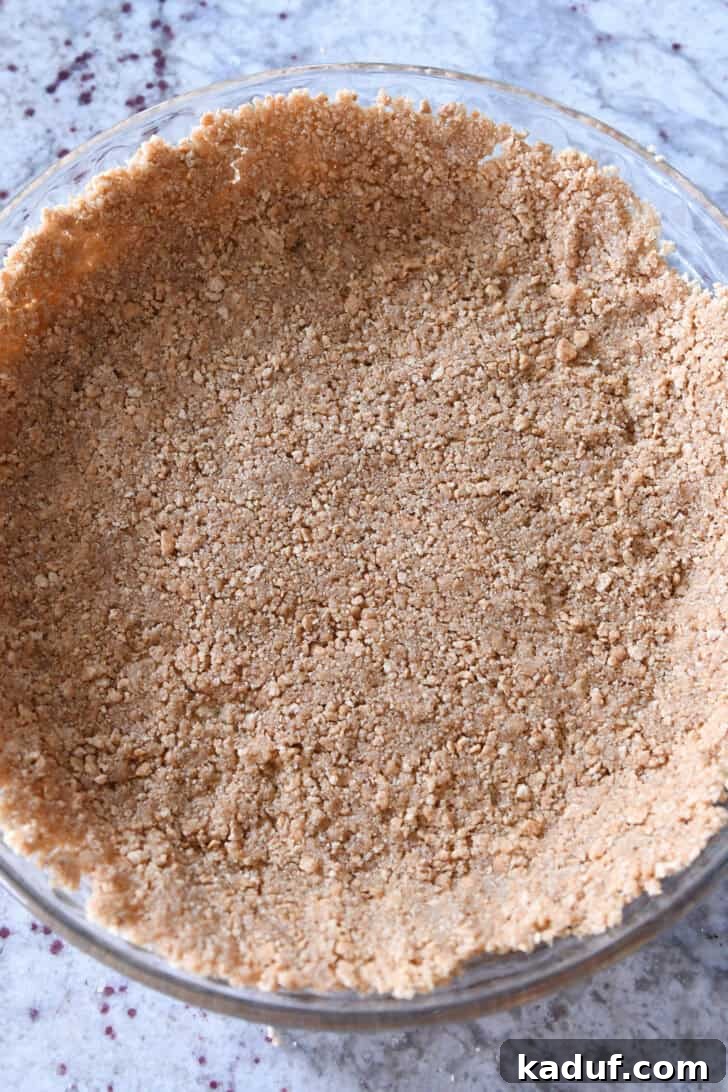 Ultimate No-Bake Cheesecake 5 Top down view of a golden brown graham cracker crust pressed evenly into a clear glass pie plate, ready for the filling.