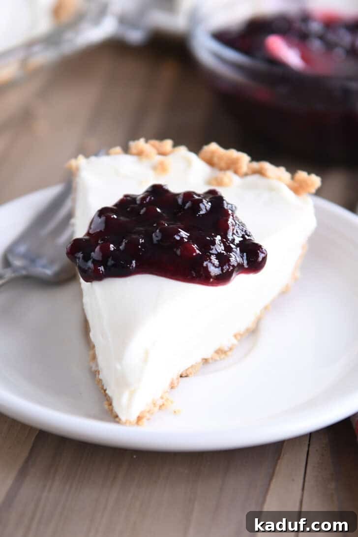 Ultimate No-Bake Cheesecake 9 A perfectly portioned slice of no-bake cheesecake with a generous dollop of rich blueberry sauce cascading over its creamy surface, presented on a simple white plate.
