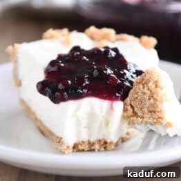 A slice of creamy no-bake cheesecake with a vibrant dollop of blueberry sauce on top, presented on a pristine white plate. A fork rests beside the slice, holding a perfect bite of the cheesecake, inviting you to taste.