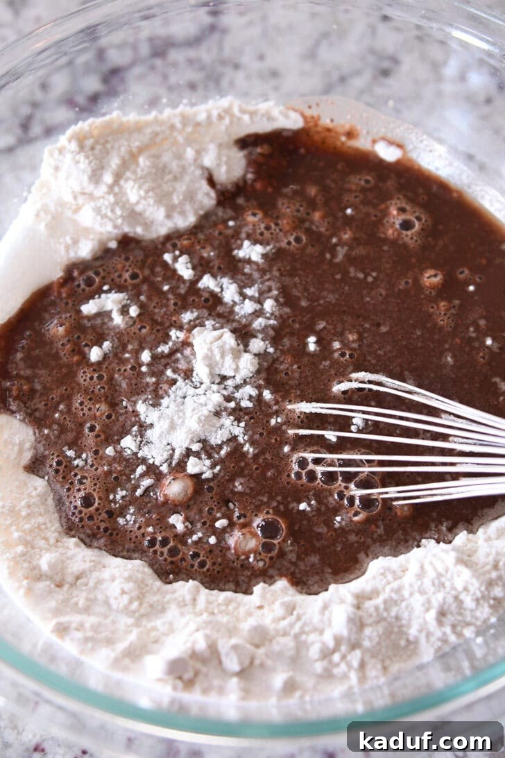 Velvet Chocolate Fudge Bundt 3 Close-up view of a whisk stirring flour and other dry ingredients into a rich chocolate cake batter in a clear glass bowl, showcasing the smooth consistency.